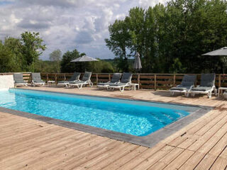 Outdoor swimming pool with clear blue water surrounded by wooden decking, lounge chairs, and umbrellas. Trees and cloudy sky in the background.