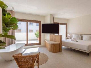 A modern hotel room with a large bed, a freestanding bathtub, a flat-screen TV mounted on a wooden partition, and a wicker chair. The room includes large windows with curtains and a view of another building.