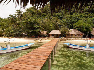 A wooden pier extends over clear water toward a tropical beach with palm trees, two small boats docked on either side, and thatched-roof buildings in the background.