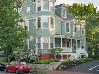 A large green three-story house with multiple windows and a porch, situated on a slightly elevated yard. A red car is parked on the street in front. Green trees surround the property.