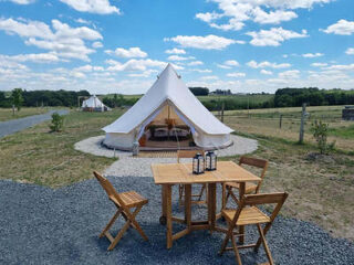 A triangular white canvas tent is set up on a grassy open field with a wooden table and three chairs outside. The sky is clear with scattered clouds.