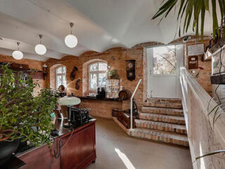 A cozy, brick-walled interior with arched windows, plants, and a staircase leading to an open door. The room is lit by round ceiling lights and furnished with a wooden desk and various decorative items.