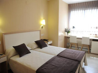 Hotel room with twin beds, white bedding with dark brown throws and pillows, a small desk with two chairs near a window, and warm-colored walls. A wall-mounted light fixture is above each bed.