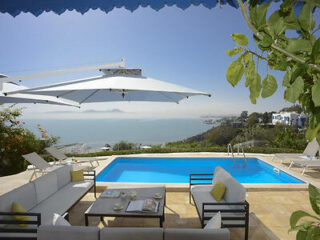 A serene poolside area overlooking a vast body of water, with white outdoor furniture and large umbrellas.