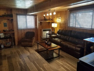 A cozy living room with wooden walls and floors, featuring a brown leather couch, an armchair, a coffee table, a small fireplace, shelves with decor, a lamp, and two windows with blinds.