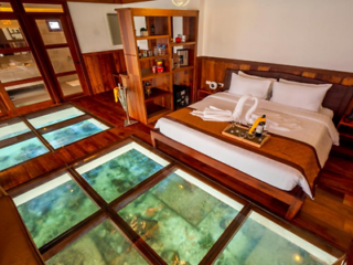 Hotel room with a double bed, wooden furnishings, a glass floor showcasing underwater views, and a small shelf divider. A folded towel swan and tray with amenities sit on the bed.