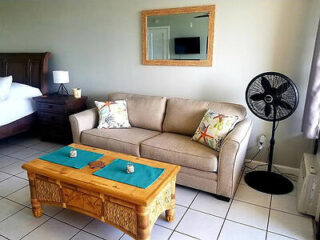 A living room with a beige sofa, two floral pillows, a wooden coffee table, a standing fan, and a wall mirror reflecting a TV. A lamp is on a side table next to the bed visible to the left.