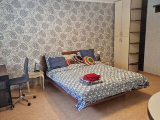 A bedroom with a patterned wallpaper, a made bed with decorative pillows, a stacked blankets, a desk with a chair, and a storage cabinet with open shelves.