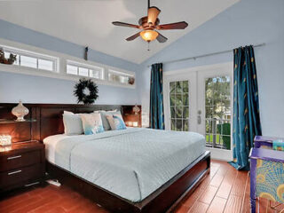 A bedroom with light blue walls, a double bed, two nightstands, a ceiling fan, windows above the bed, and French doors leading outside.