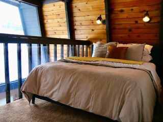 A bed with beige sheets, brown pillows, and a yellow throw is placed in a cozy room with wooden walls, two wall-mounted lights, and a railing overlooking a window.