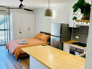 A studio apartment featuring a bed with orange bedding, a kitchen area with a fridge, oven, and cabinets, a ceiling fan, and a green plant on a shelf.