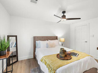 A bedroom with a double bed, tufted headboard, yellow throw blanket, side table with lamp, ceiling fan, wall mirror, and small potted plant on another table.