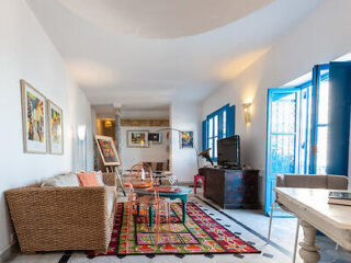 A brightly lit living room with wicker furniture, colorful decor, and blue-framed windows opening to the outside.