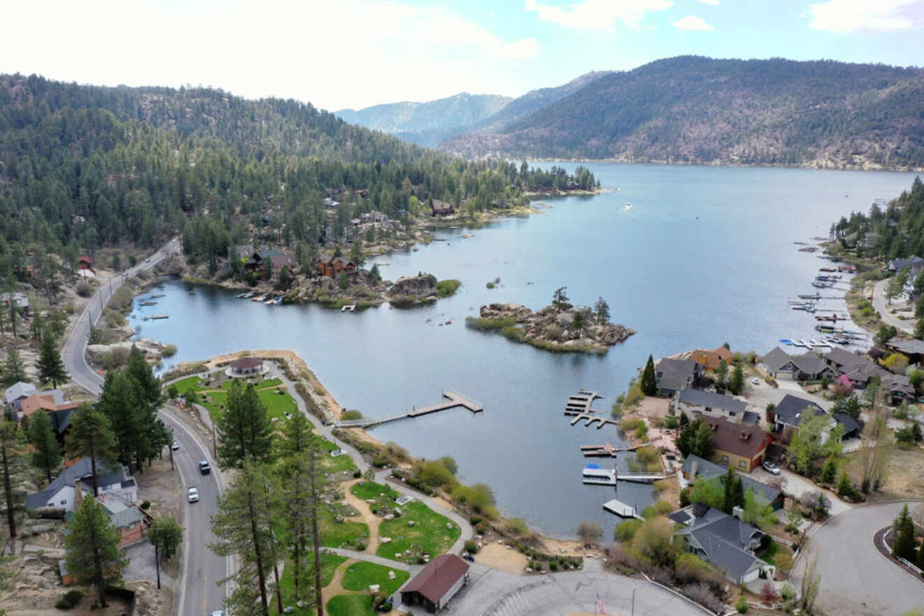 Aerial view of a serene lakeside community with houses, docks, a winding road, green trees, and hills in the background.