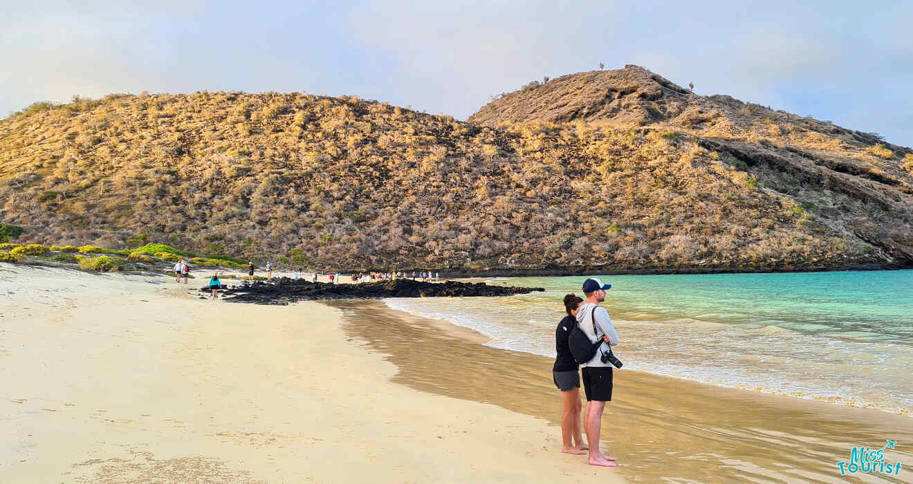 A couple stands on a sandy beach facing the water, with a rocky hill in the background and several other people further down the shore.