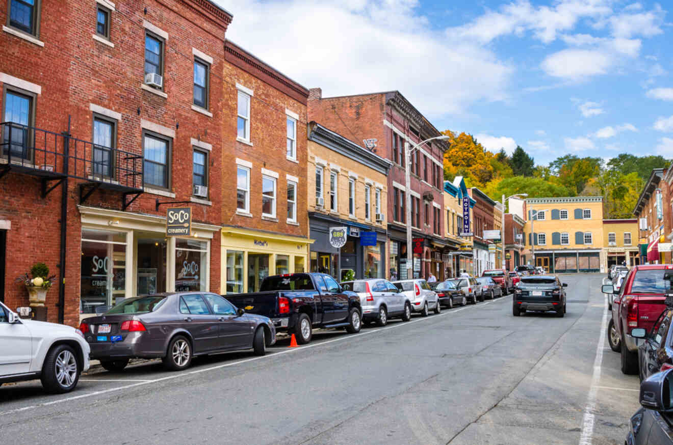 A street lined with parked cars and various brick buildings housing shops and businesses under a partly cloudy sky.