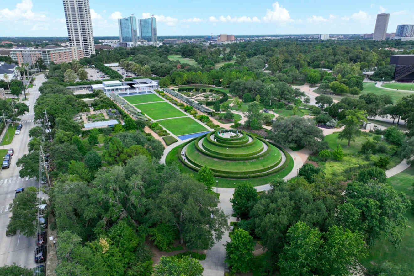 Aerial view of a green park with a tiered circular garden, rectangular pond, and adjacent modern buildings. Trees and vegetation surround the area.