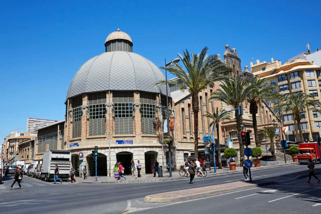 Street corner with a large domed building, palm trees, pedestrians, and traffic signals under a clear blue sky.