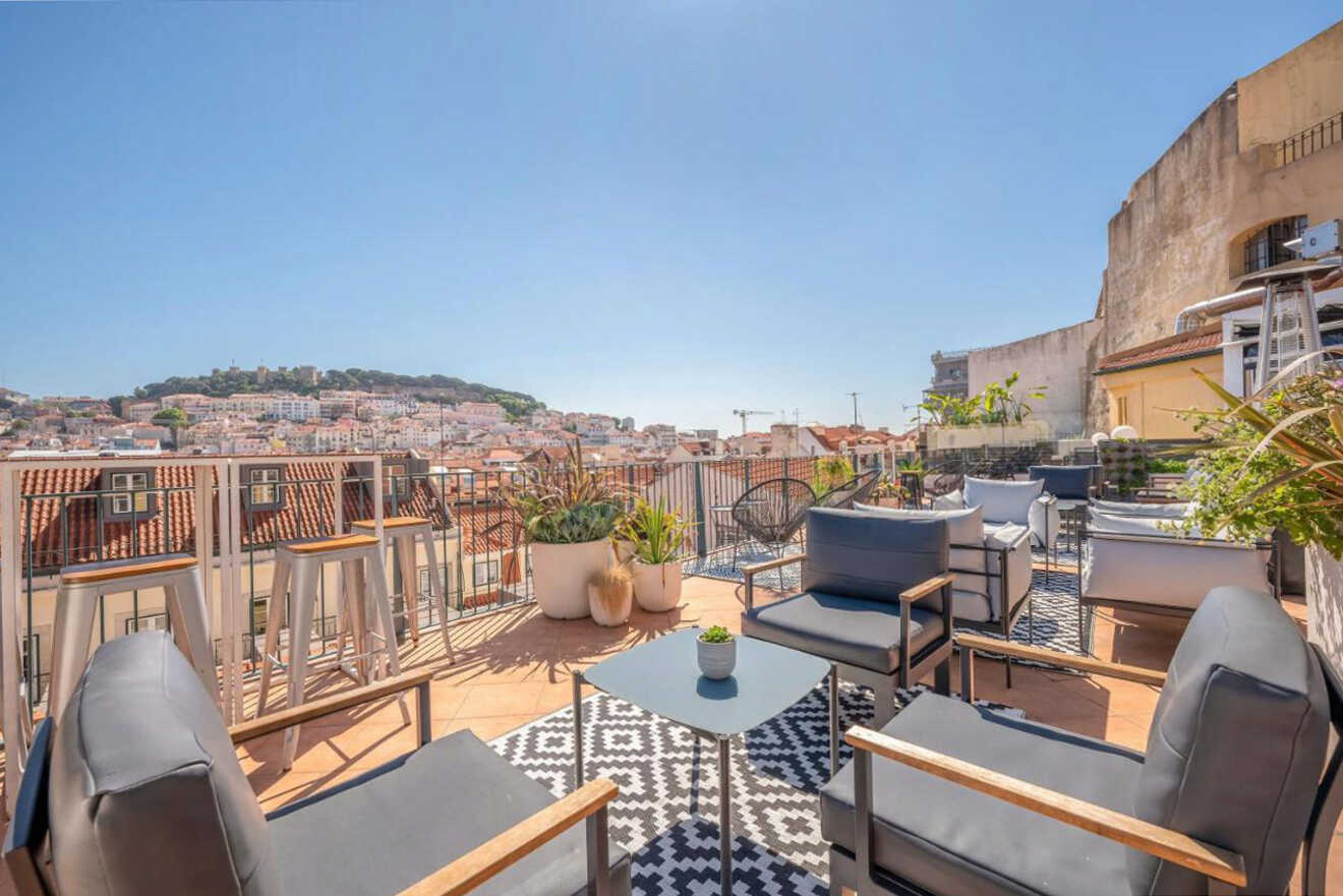 Rooftop terrace with modern furniture, plants, and a patterned rug, offering a view of a hilly cityscape under a clear blue sky.