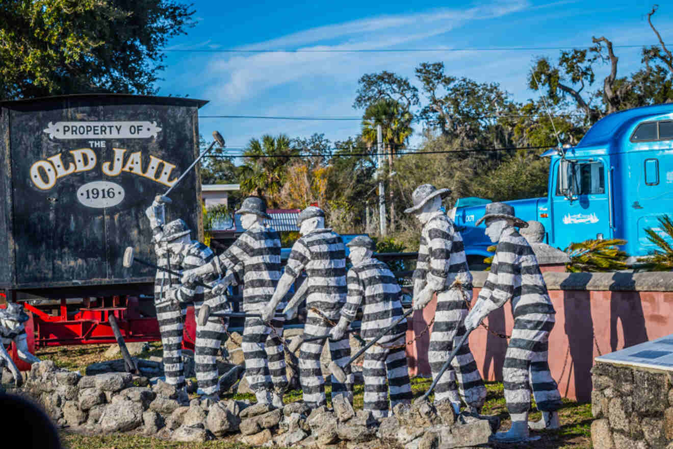 A sculpture of prisoners in striped uniforms doing manual labor is displayed outdoors, positioned next to a sign that reads "Property of The Old Jail 1916." The background features trees and a blue truck.