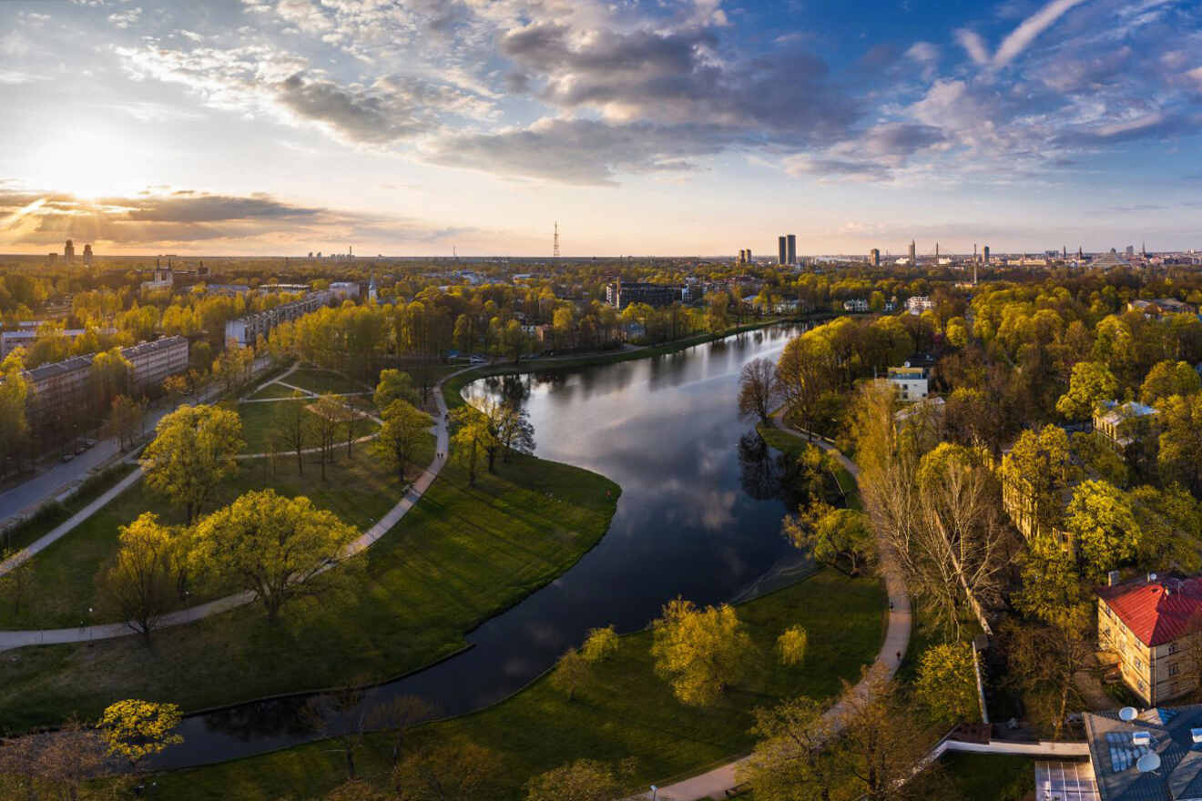 Aerial view of a city park with a winding river, surrounded by trees and grassy areas, under a partly cloudy sky during sunset. Buildings and cityscape are visible in the background.