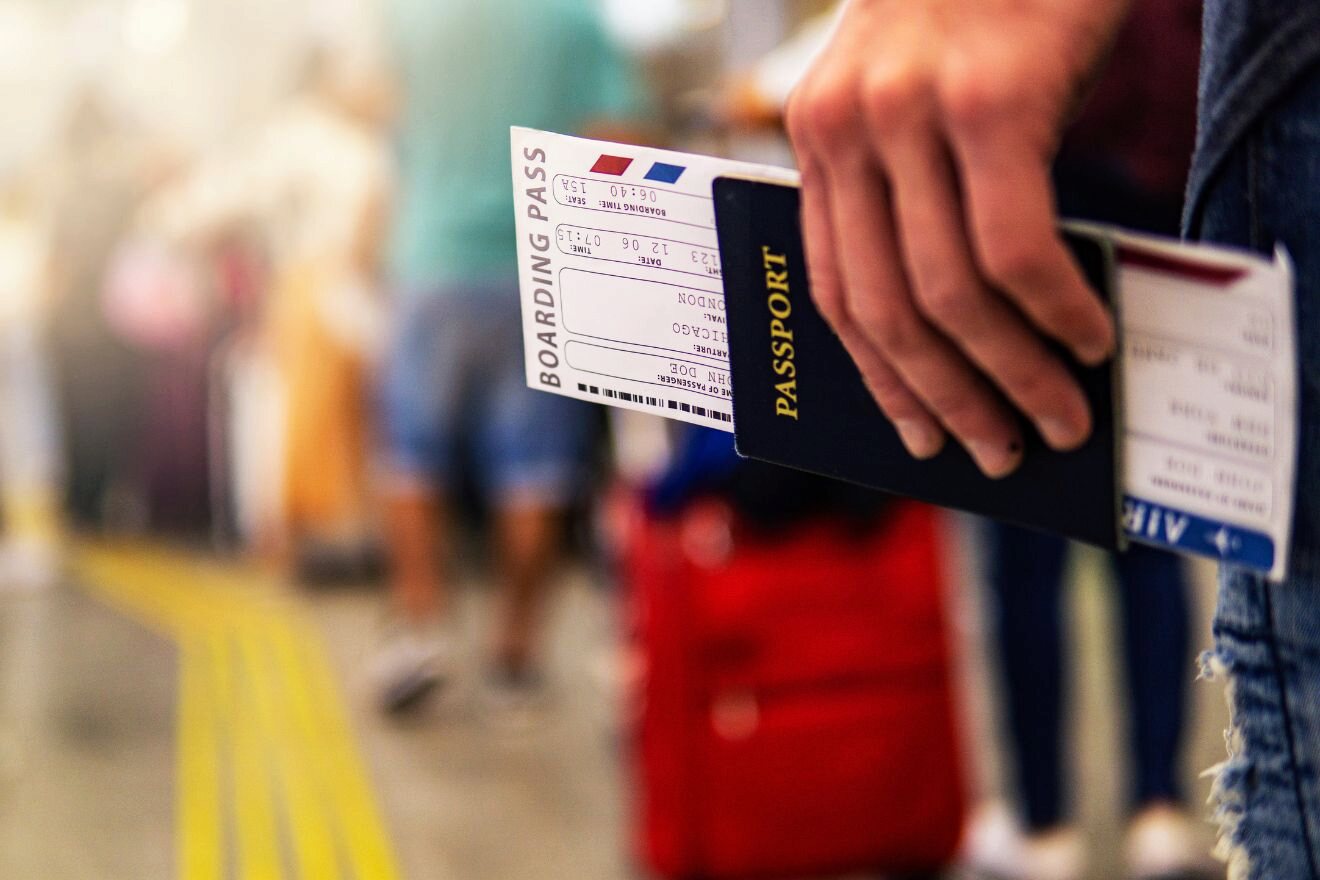 Close-up of a hand holding a passport with a boarding pass, standing in a line at an airport. The background shows blurred travelers and luggage.