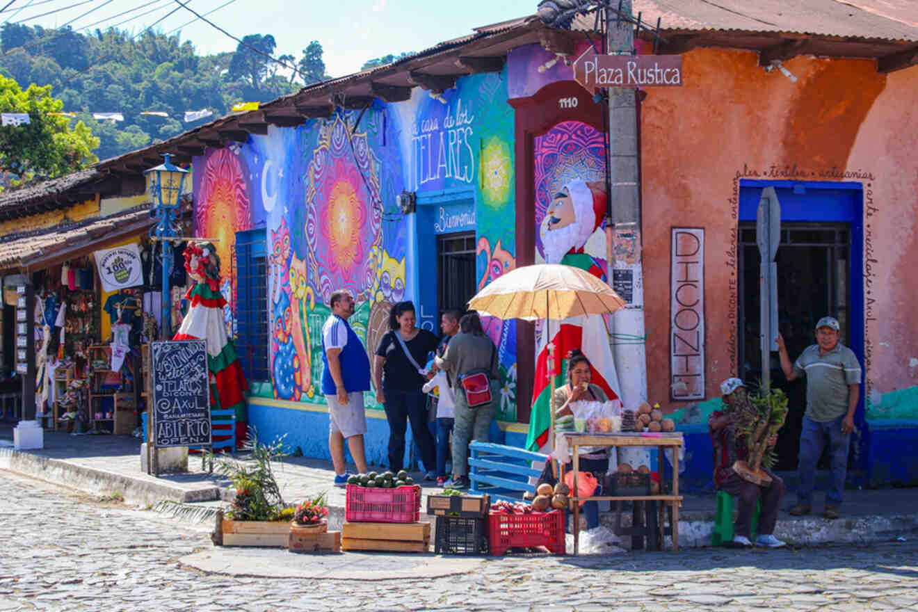 People gather outside colorful, mural-decorated buildings on a cobblestone street. A vendor sells produce under an umbrella, and festive decorations, including a Santa figure, adorn the area.