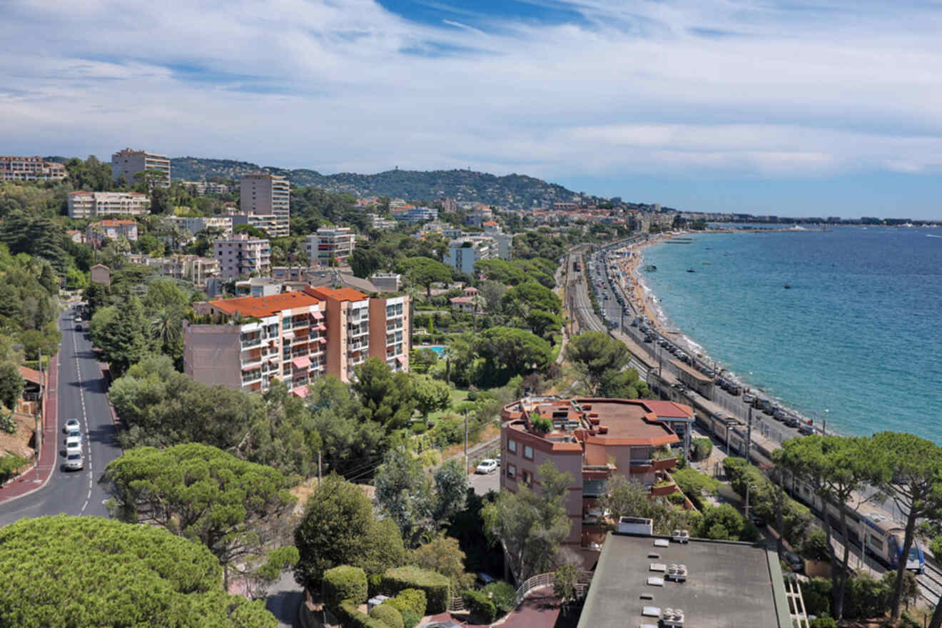 A coastal cityscape with residential buildings, a road, and a beach along the shoreline. Trees and greenery are visible among the buildings, and a clear blue sky can be seen above.