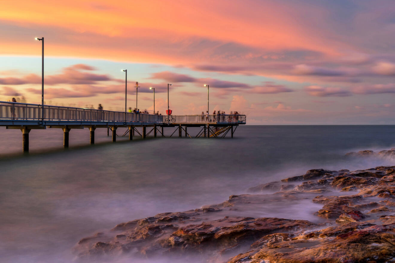 A long pier extends over the ocean at sunset, with people standing at its end. The sky is a mix of orange and pink hues, and waves gently splash against rocks in the foreground.