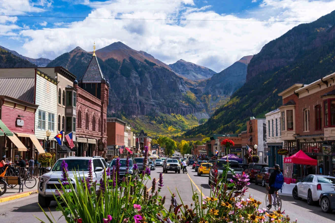 A vibrant street in a mountain town with colorful buildings, people walking, and cars driving. Mountains and a cloudy sky are visible in the background. Flower planters line the street in the foreground.