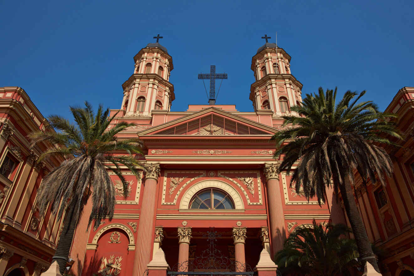 The façade of a historic church in Santiago, Chile, featuring red walls, intricate detailing, and tall palm trees in the foreground.