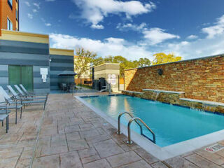 Outdoor pool area with lounge chairs, an umbrella-shaded seating area, and a stone wall fountain feature. The sky is clear with some clouds.