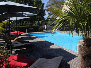 Outdoor swimming pool with lounge chairs and umbrellas in a shaded area, surrounded by greenery and palm trees.