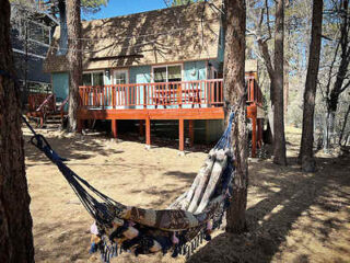 A hammock hangs between trees in front of a wooden house with a large elevated deck.