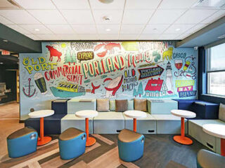 A colorful mural in a modern cafe or lounge area depicts various Portland, Maine landmarks and phrases. The space includes white round tables, orange table bases, and a mix of seating options.