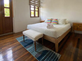 Bright bedroom featuring a wooden bed with white linens and colorful accent pillows, along with hardwood floors.