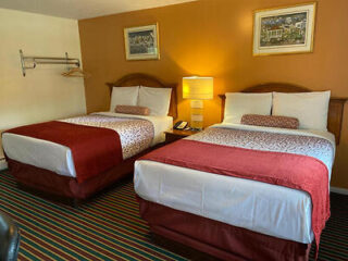 A hotel room with two made-up queen-sized beds, each with white and red bedding, a nightstand with a lamp between them, and framed pictures on the wall. Carpet is striped, and there's a clothes rack.