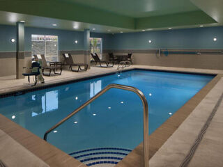 Indoor swimming pool with metal handrails, surrounded by lounge chairs and tables, in a well-lit room with light blue walls and ceiling.