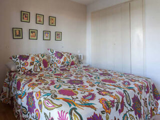 A bedroom with a double bed covered in a floral-patterned bedspread and matching pillows. There are framed pictures on the wall and a closed wardrobe in the background.