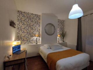 A bedroom with a double bed, white bedding, and an orange throw blanket. The room is decorated with patterned wallpaper, a round mirror, and a work desk with a chair and laptop. A pendant light hangs above.