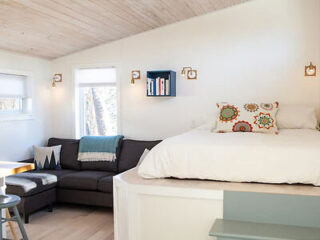 A small, well-lit bedroom features a raised bed with patterned pillows, a sectional sofa with a blanket, a small table with stools, and a shelf with books on a white wall.