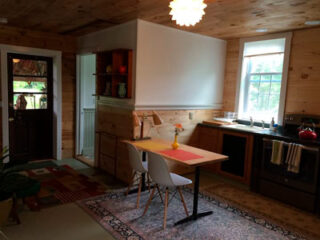 A small wooden kitchen with a dining table, two chairs, a door to the left, cabinets, a stove, a sink, a window, and a hanging light fixture. A vase with flowers is on the table.