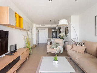 A modern living room with a beige couch, armchair, white coffee table, wall-mounted TV, and shelves. The space is well-lit with ample natural light, leading to a dining area and kitchen in the background.