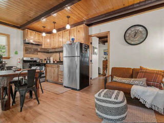 A cozy kitchen and living area with wooden ceiling, stainless steel fridge, dining table with chairs, brown sofa with plaid blanket and "welcome home" pillow, wall clock, and patterned ottoman.