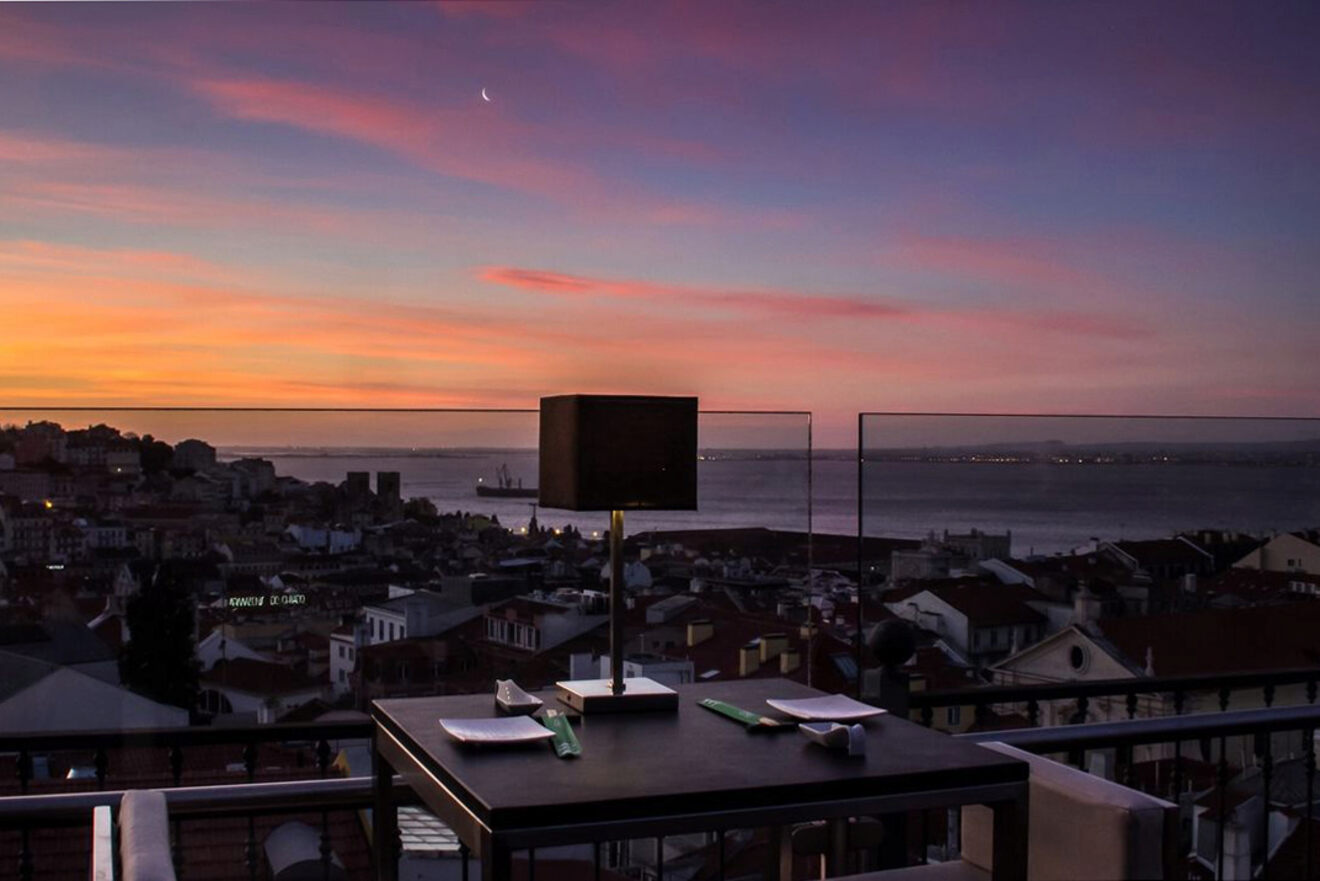 A rooftop dining table set with a lamp and place settings overlooks a cityscape at dusk, with a moon and colorful sunset sky in the background.