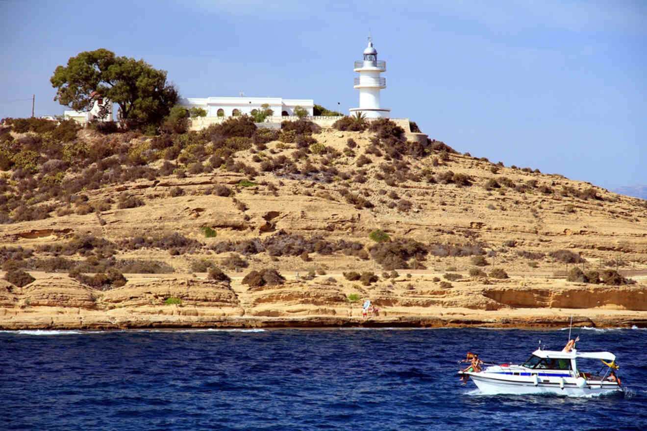 A lighthouse on a rocky hilltop with a building beside it near the shoreline, and a small boat with people on the water in the foreground.