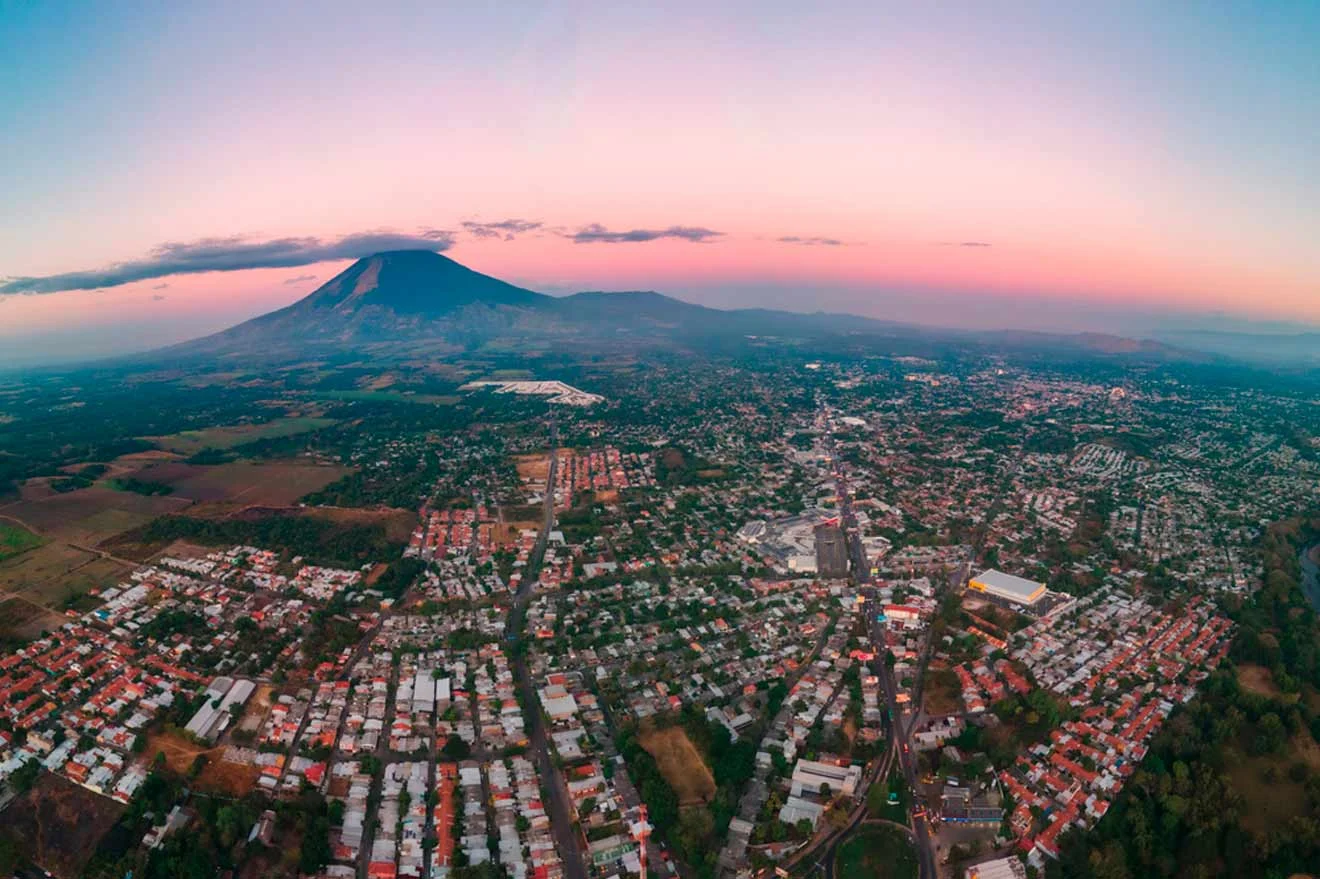 Aerial view of a city with a sprawling residential area in the foreground, and a large volcanic mountain under a pink and orange sky in the background.