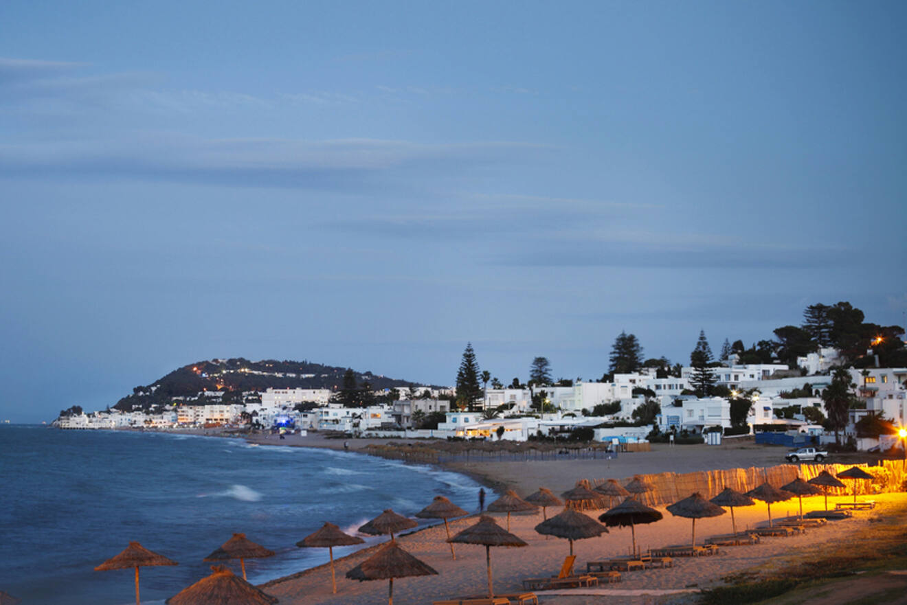 A quiet evening view of a beach in Gammarth, with rows of straw umbrellas and white buildings illuminated in the distance.