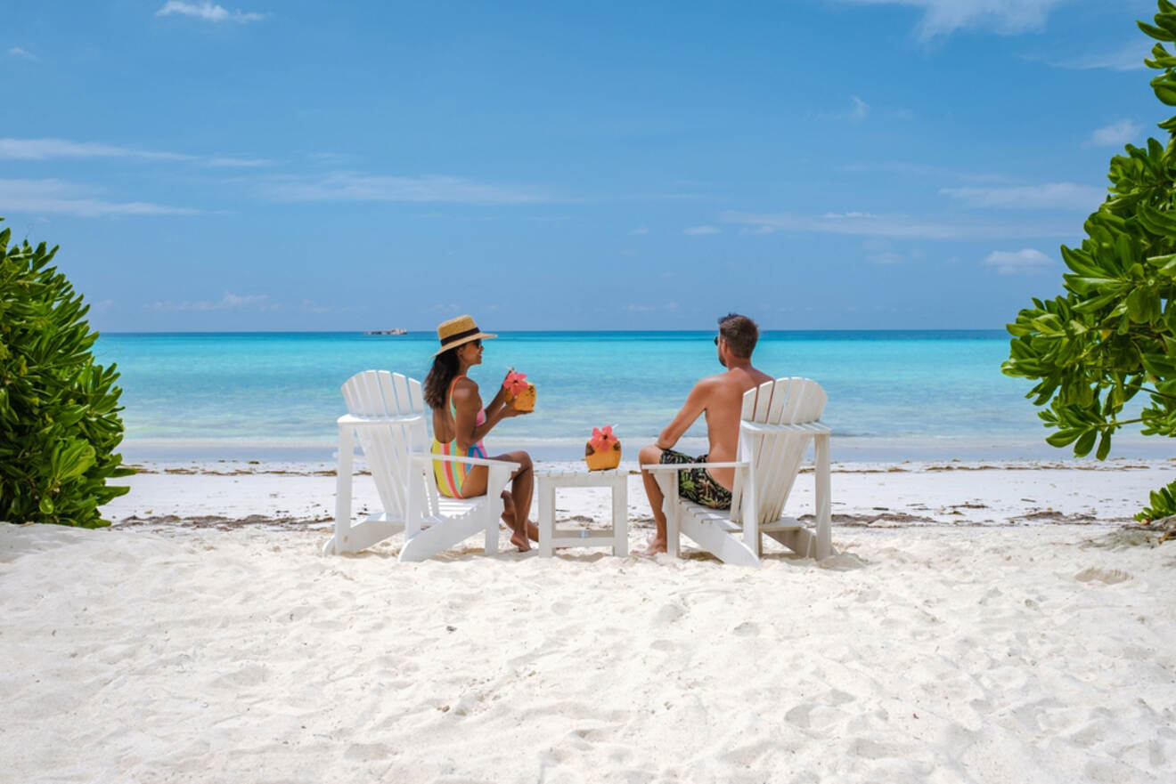 A couple sitting on white lounge chairs on a sandy beach, enjoying tropical drinks and the view of the turquoise ocean under a clear blue sky.