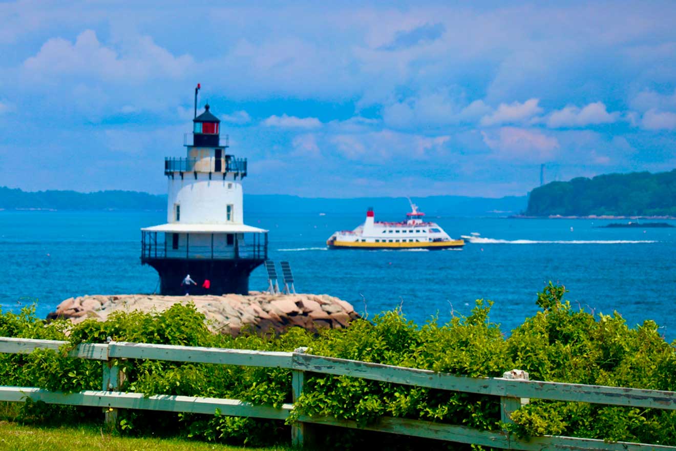 A white lighthouse stands on rocky terrain by the water as a ferry boat with a yellow bottom and red upper section sails in the background. Green foliage and a wooden fence are in the foreground.